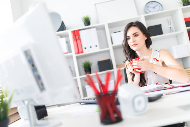 Die schöne junge Frau sitzt am Schreibtisch, blickt auf den Computerbildschirm und hält einen roten Becher in der Hand. - Foto, Bild