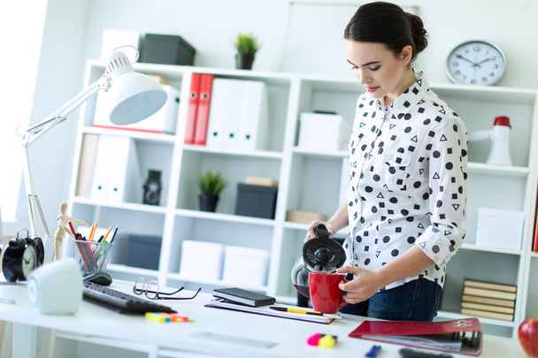 Egy fiatal lány az irodában a tábla közelében áll, és önti a kávét a kávéfőző a piros pohár. - Fotó, kép