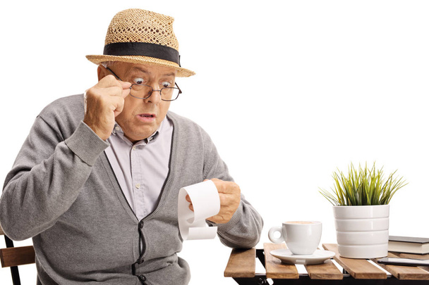 Shocked mature man seated at a coffee table looking at the bill isolated on white background - Foto, Bild