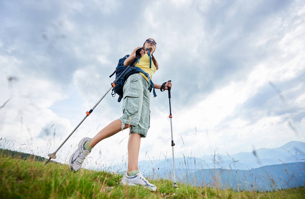 Low angle view of sporty woman tourist hiking mountain trail, walking on grassy hill, wearing backpack and sunglasses, using trekking sticks, enjoying summer day. Outdoor activity, lifestyle concept - Foto, afbeelding