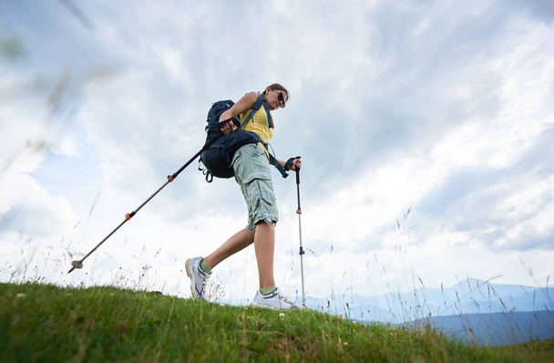 Low angle view of attractive woman tourist hiking in Carpathian mountain trail, walking on grassy hill, wearing backpack and sunglasses, using trekking sticks, enjoying summer day. Lifestyle concept - Photo, image
