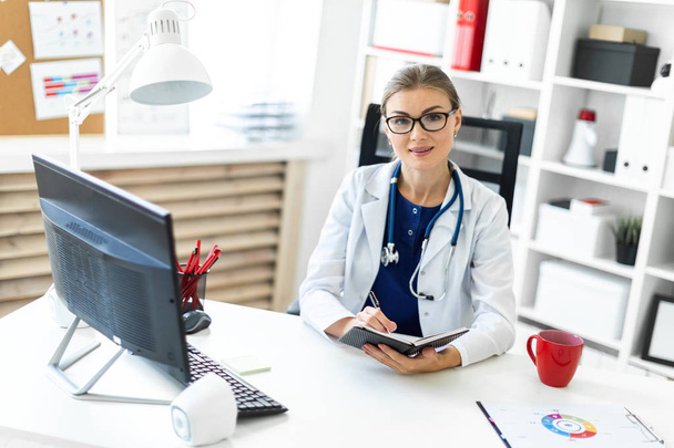 A young girl in a white robe sits at a table in the office and holds a pen and a notebook in her hand. A stethoscope hangs around her neck. - Photo, Image