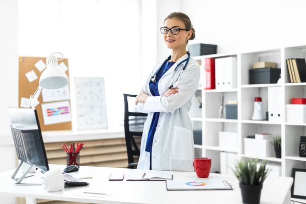 Młoda dziewczyna w biały płaszcz jest stojący w pobliżu tabeli w jej biurze. Stetoskop zawiesza się wokół szyi. - Zdjęcie, obraz