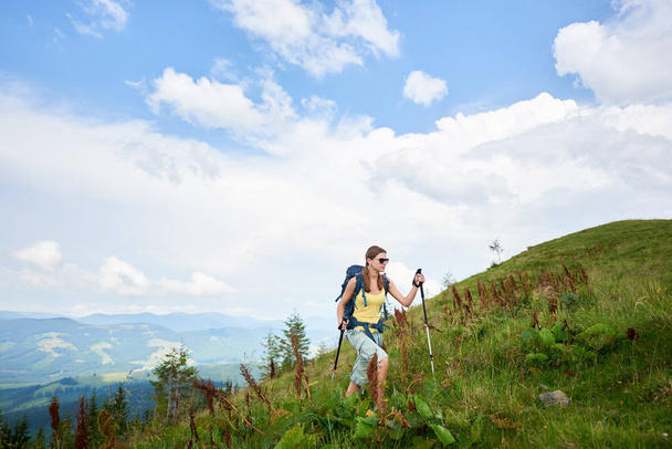 Atraente mulher turista caminhadas trilha de montanha, andando em colina gramada, vestindo mochila e óculos de sol, usando paus de trekking, aproveitando o dia de verão. Atividade ao ar livre, conceito de estilo de vida
 - Foto, Imagem