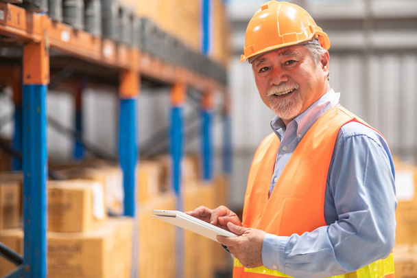 Senior warehouse operator portrait. Old chinese man in safety hat pose smile holding tablet with bottle warehouse in background. - Foto, afbeelding