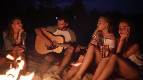 Beach party při západu slunce s ohněm. Přátelé, seděli kolem ohně, popíjeli pivo a zpívá na kytaru. Muži a ženy drží skleněných lahví nápoj singalong, vousatý chlap hraje na kytaru. - Záběry, video