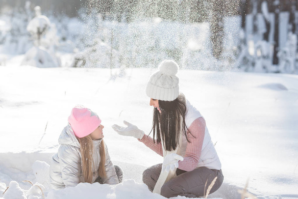 Young pretty mother and her little girl having fun outdoors in the winter. Cheerful child and her beautiful mom playing with snow at the nature. Blowing snow - Photo, Image