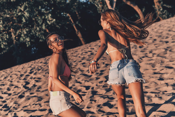 happy young women in shorts and swimwear bras running on sandy beach - Photo, image