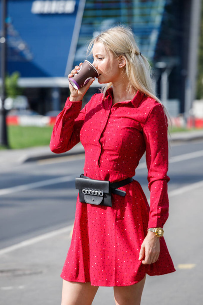 Outdoor fashion portrait of beautiful blond woman in red dress holding coffee cup, street background  - Foto, imagen