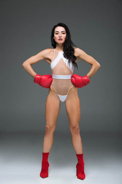 Sexy sportive woman in white swimwear and boxing gloves standing with hands akimbo, on grey - Фото, зображення