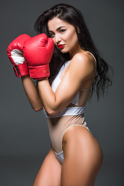 vista lateral de mujer deportiva sexy en ropa deportiva blanca y guantes de boxeo mirando a la cámara aislada en gris
 - Foto, Imagen