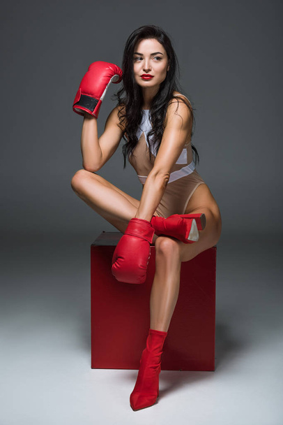 sexy sportive woman in white swimwear and boxing gloves sitting on red cube and looking away on grey - Photo, Image