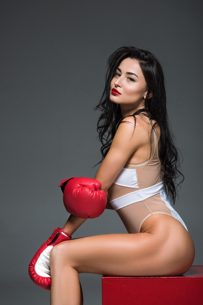 vista laterale di sexy donna sportiva in costume da bagno bianco e guanti da boxe seduti sul cubo rosso isolato sul grigio
  - Foto, immagini