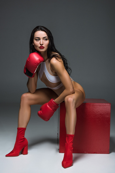 sexy sportive woman in white swimwear and boxing gloves sitting on red cube on grey  - Foto, Bild