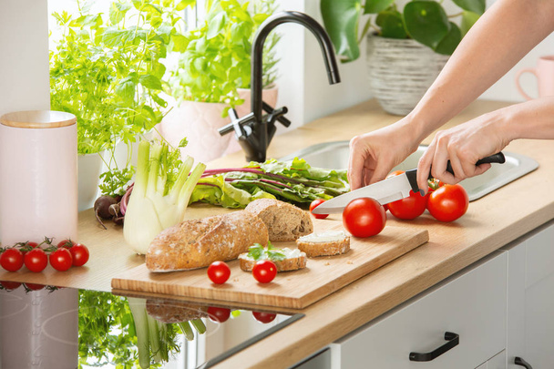 Woman preparing healthy breakfast, cutting a tomato in half and organic herbs and vegetables in a sunny kitchen interior - Photo, Image