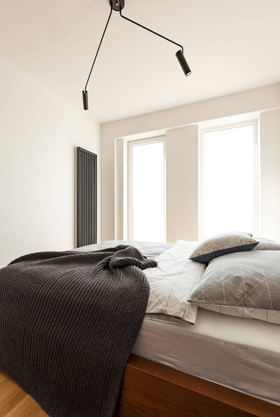 Ángulo lateral de una cama doble con manta y almohadas, ventana grande y lámpara negra en el interior de un dormitorio
 - Foto, Imagen