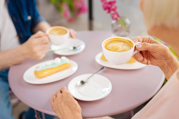 Tender crema. Close up of coffee hold by female hands while eclairs waiting on surface - Foto, afbeelding