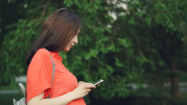 Side view of attractive pregnant woman walking in park and using smart phone, person is watching screen then looking around enjoying beautiful nature. - Séquence, vidéo