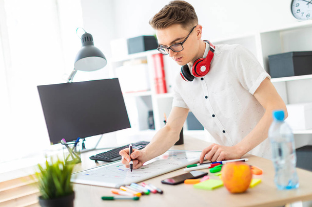 Молодой человек в очках стоит возле компьютерного стола. Молодой человек рисует маркер на магнитной доске. На шее висят наушники.
. - Фото, изображение