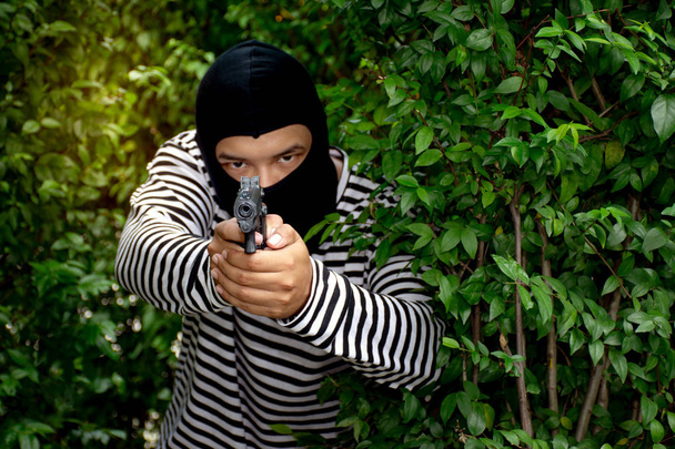 Los terroristas están detrás del arbusto apuntando su arma bajo la máscara con la cara borrosa detrás
 - Foto, Imagen