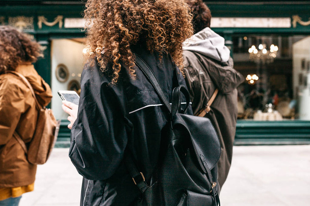 Egy turista lány göndör haja egy hátizsákban vagy a diák, az utcán, és egy mozgatható telefon vagy mobiltelefon segítségével. - Fotó, kép