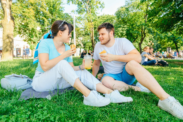 couple sitting on green grass eating burgers drinking smoothie. wide angel - Foto, imagen