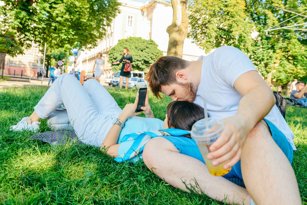 couple resting on grass in city park. drinking smoothie surfing phone - Foto, afbeelding