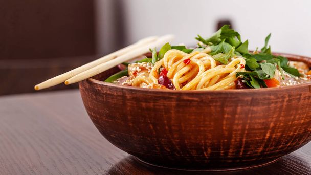 Asian Oriental Cuisine. Japanese noodles with chicken and vegetables, sesame and parsley, in a plate of red clay, stands on a table in a restaurant. Copy space, selective focus - Photo, image