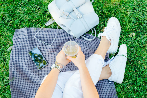 woman holding cup with cool drink while sitting on blanket on grass - Foto, Imagen