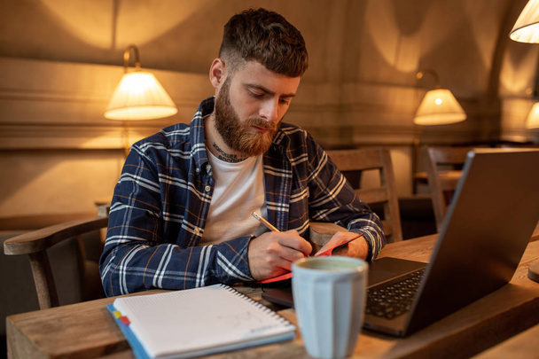 Случайный бизнесмен или фрилансер, планирующий свою работу над ноутбуком, работающий на ноутбуке со смартфоном, чашкой кофе на столе в кафе или домашнем офисе
. - Фото, изображение