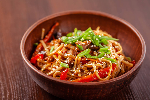 Asian Oriental Cuisine. Japanese noodles with chicken and vegetables, sesame and parsley, in a plate of red clay, stands on a table in a restaurant. Copy space, selective focus - Zdjęcie, obraz