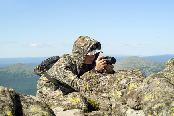 girl wildlife photographer in camouflage suit in the mountains hiding behind a stone photographing something - Photo, Image
