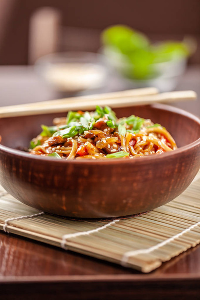 Asian Oriental Cuisine. Japanese noodles with chicken and vegetables, sesame and parsley, in a plate of red clay, stands on a table in a restaurant. Copy space, selective focus - Fotografie, Obrázek