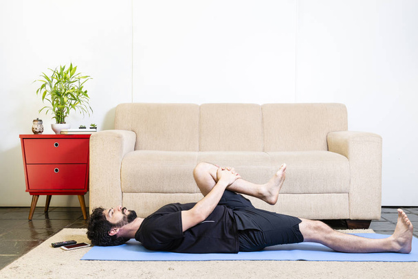 Beautiful caucasian bearded man in black clothes on blue yogamat doing half wind-relieving pose - Photo, image