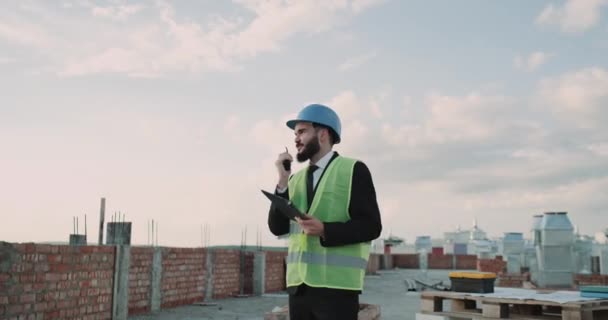 On rooftop at construction site businessman wearing a safety helmet and high visibility vest speaking using a black radio. - Video, Çekim
