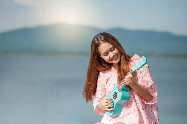 asian lady with hat play ukulele bossanova music in summer time.Young cute woman playing music outdoors.A happy young girl enjoys playing ukulele under a lake. Beautiful nature in the background. - Photo, image