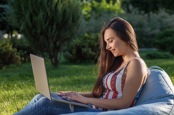 Молодая симпатичная девушка с ноутбуком, сидит на фасолевом пакете в саду или парке, на зеленой траве. Концепция покупок
 - Фото, изображение