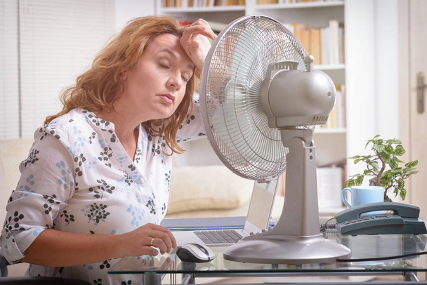 Woman suffers from heat while working in the office and tries to cool off by the fan - Zdjęcie, obraz
