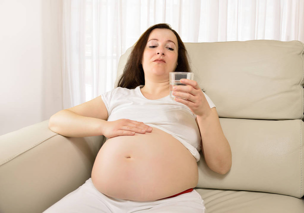An pregnant woman sitting in the live room thinking and holding a glass of water - Foto, imagen
