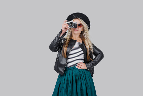 Fashion look, happy cool young woman model with retro film camera makes a photo. wearing an elegant hat, black jacket, blondy hair outdoors over grey background - Fotografie, Obrázek