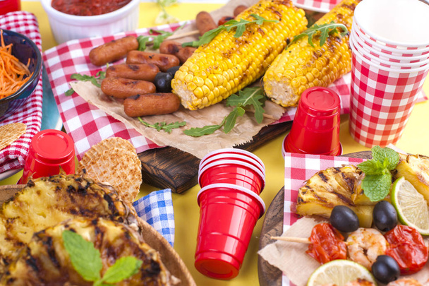 Picnic with grilled food. Sausages and corn on barbecue, shrimp, vegetables and fruits. Delicious summer lunch and plastic dishes. Top view. - Foto, Imagem