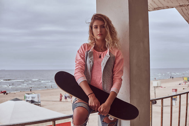 Porträt eines charmanten jungen Mädchens mit blonden Haaren, rosa Jacke und zerrissenen Jeans hält ein Skateboard in der Hand, während es auf einer Leitplanke vor der Küste sitzt. - Foto, Bild