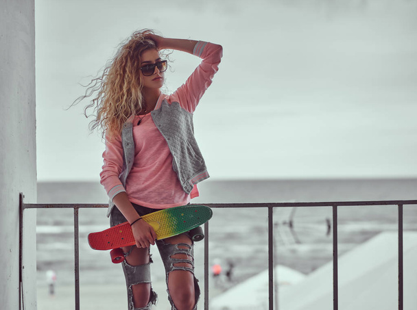 Retrato de uma jovem sensual com cabelo loiro em óculos de sol vestidos com uma jaqueta rosa em pé perto de um corrimão contra fundo da costa do mar preto e branco
. - Foto, Imagem
