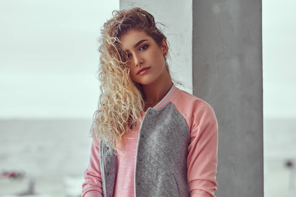 Close-up portrait of sensual young girl with curly blonde hair dressed in a pink t-shirt and jacket posing against black and white sea coast background. - Foto, afbeelding