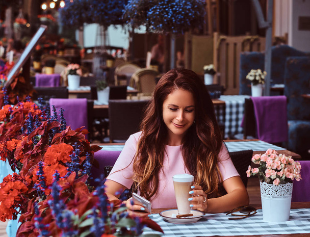 Feliz mujer de negocios de mediana edad con el pelo largo marrón con un vestido rosa sostiene el teléfono inteligente mientras está sentado en el café al aire libre
. - Foto, imagen