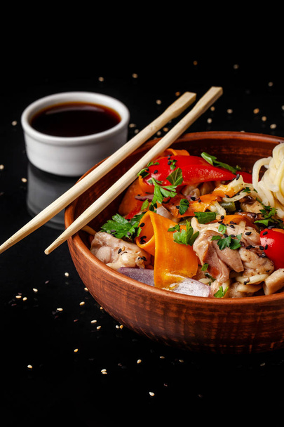 Japanese or Chinese udon noodles with chicken and vegetables, balkar pepper, carrots, parsley, white and black sesame, red onion. noodles in a clay red plate, next to soy sauce and sticks - Fotografie, Obrázek