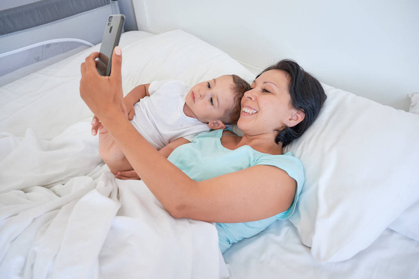 mom and baby lie together in bed and take selfies - Foto, afbeelding