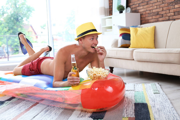 Shirtless man with inflatable mattress, drink and popcorn on floor at home - Photo, image