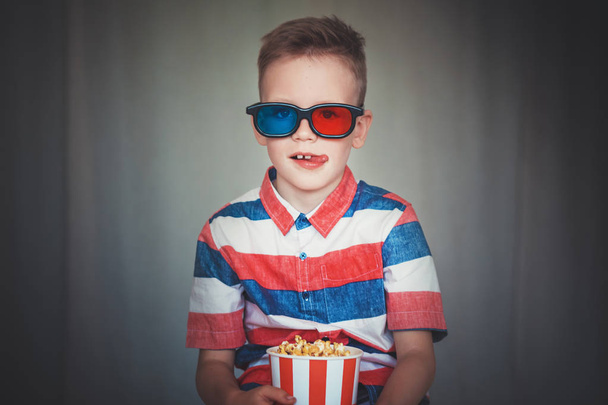 Young boy watch a movie in 3D glasses at the cinema or at home. Little kid eat popcorn over gray background. Home theater. Cute Child in vintage cinema eyeglasses. Entertainment concept.  - Photo, Image