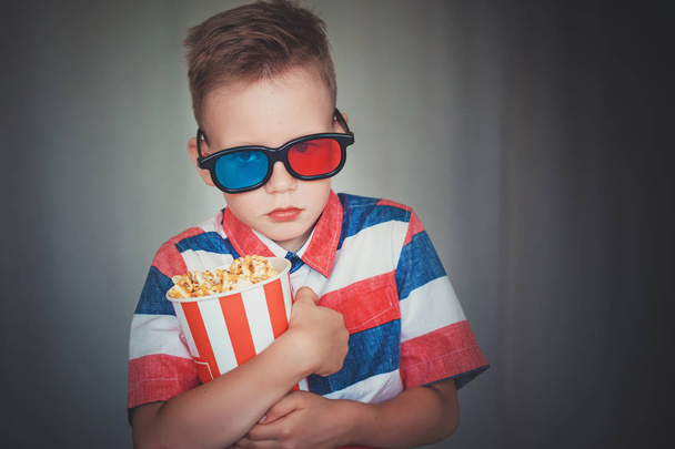 Young boy watch a movie in 3D glasses at the cinema or at home. Little kid eat popcorn over gray background. Home theater. Cute Child in vintage cinema eyeglasses. Entertainment concept.  - Foto, Bild
